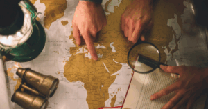 Imagem mostrando duas mãos em apontando a América no mapa e uma das mãos está segurando uma lupa.